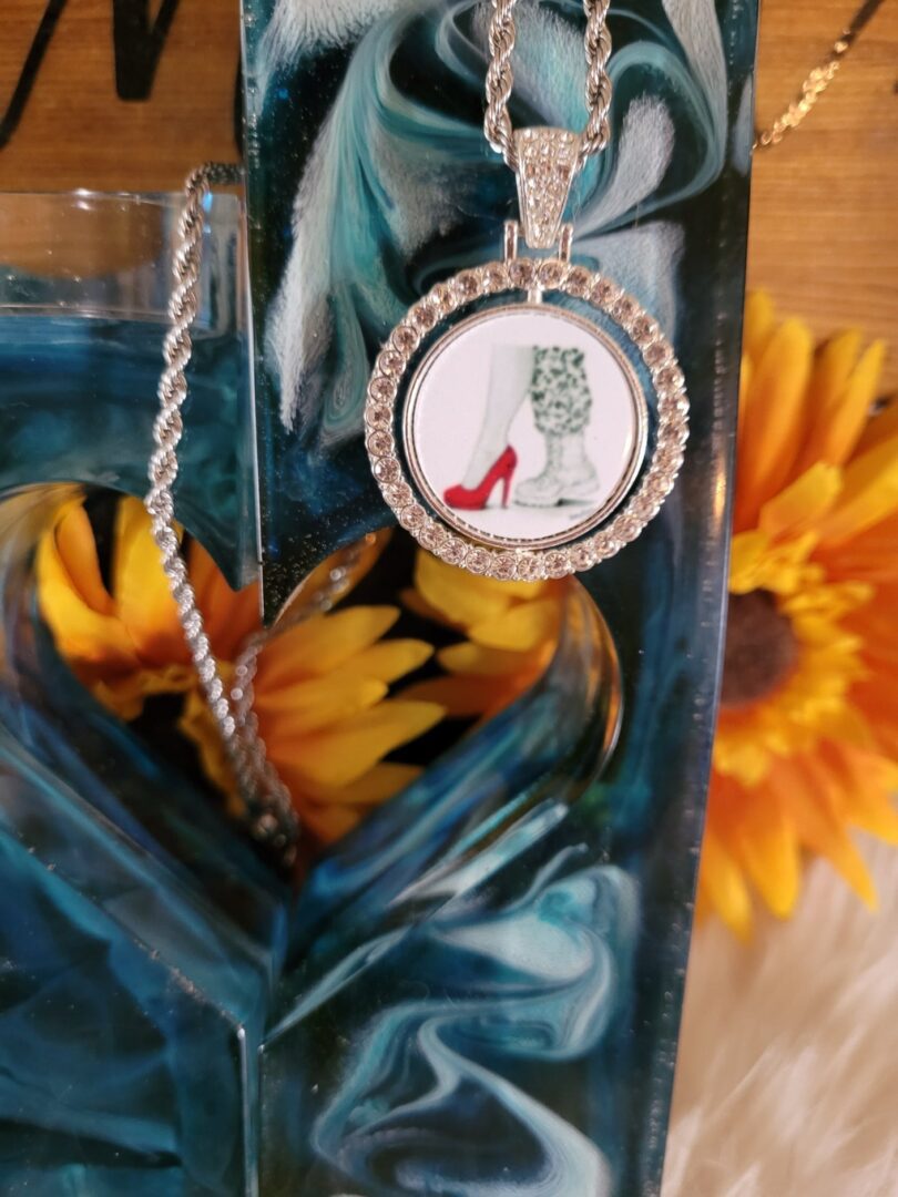 A close-up of a 24 in Stainless Steel Kelly McCook Series Diamond Necklace with a design of red high-heeled shoes, surrounded by crystals, hanging against a blue and white swirled background with sunflowers at the base.