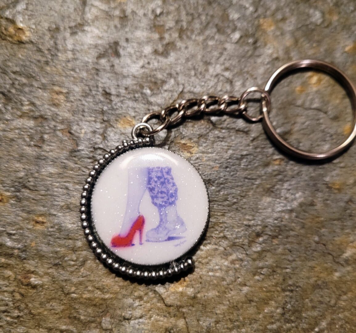 Kelly McCook Epoxy Inlay Keychain with an image of a red high heel and blue patterned boot against a white background, attached to a metal ring, on a textured surface.
