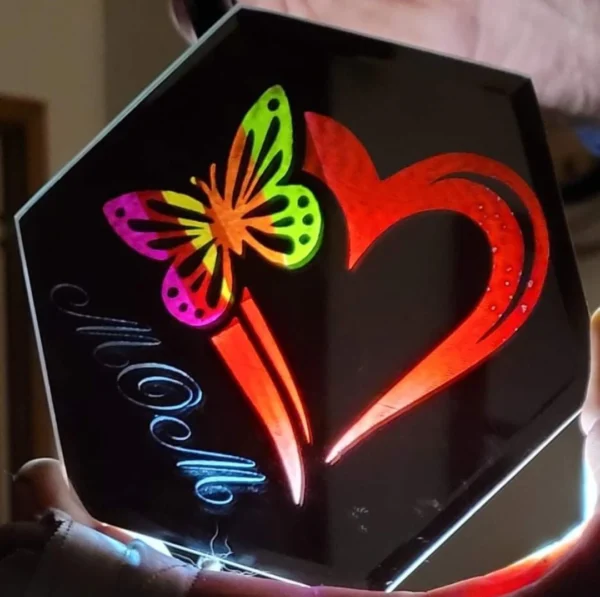 A Custom Engraved Mirror held up to the light, featuring a colorful butterfly design and a red heart, with the name "noelle" written in cursive.