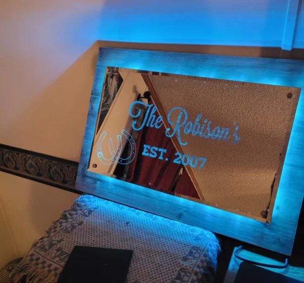 Custom Engraved Mirrors reading "the robison's est. 2007" with blue backlighting, mounted on a wooden frame above a headboard.