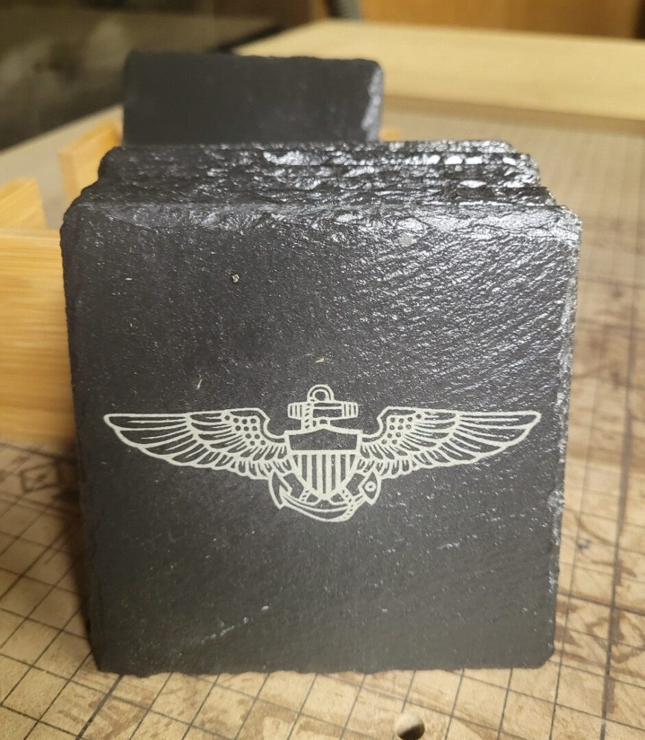 Sentence with Product Name: Custom Engraved Coasters with a black slate coaster featuring a white engraved emblem of wings and an anchor, resting on a wooden table with a visible grid background.