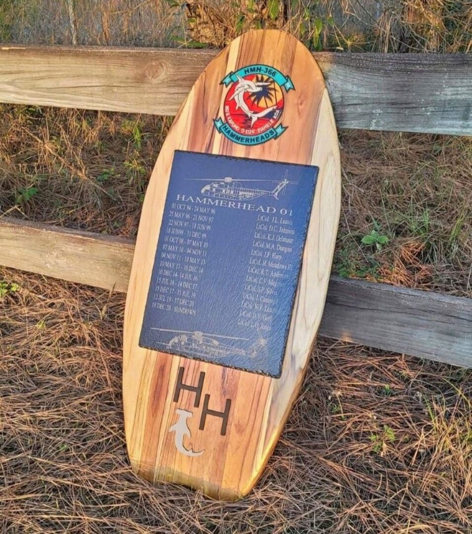 Wooden plaque mounted on a surfboard, displaying detailed text and logos, placed on a rustic bench with a background of dry grass.