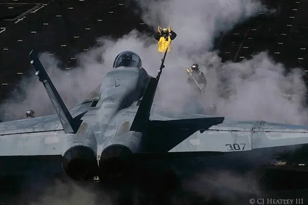 A F-18 fighter jet releases steam on an aircraft carrier as a crew member in a yellow suit directs the pilot.