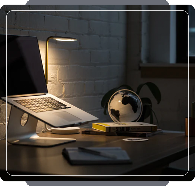 A dimly lit office with an open laptop, a desk lamp, books, and a globe on a table, against a white brick wall.